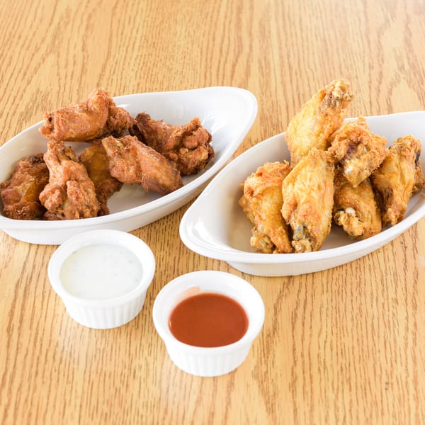 Two white dishes filled with crispy fried chicken wings, with small cups of ranch and hot sauce.