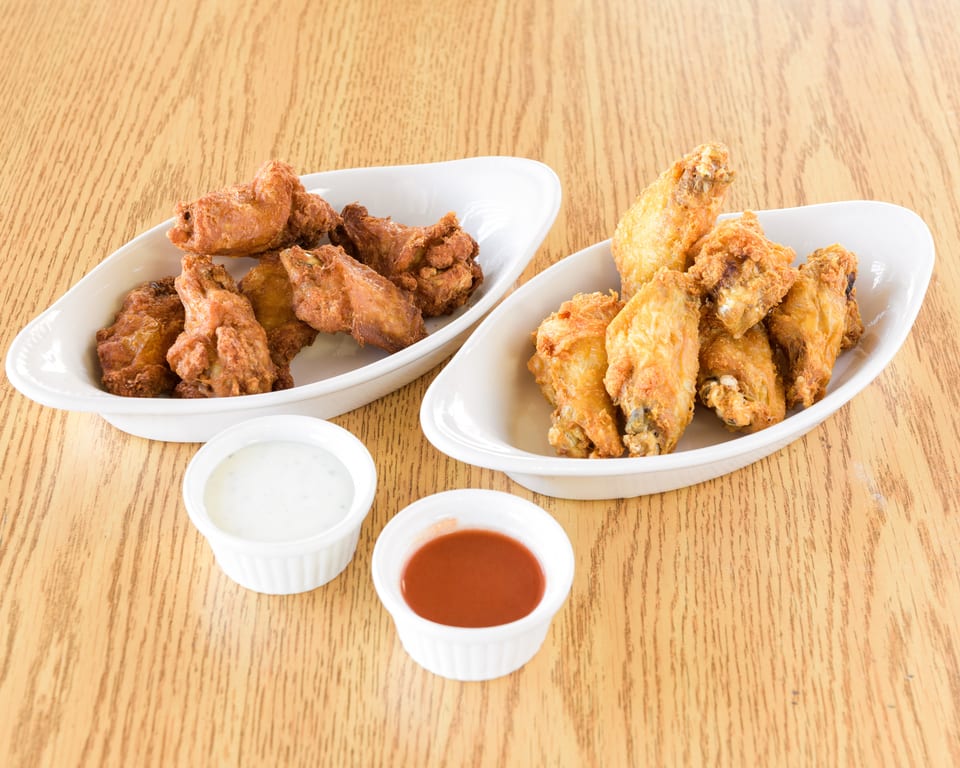 Two white dishes filled with crispy fried chicken wings, with small cups of ranch and hot sauce.