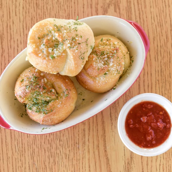 Three golden-brown garlic knots with herbs and parmesan in a dish, with a side of marinara sauce.