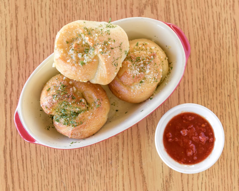 Three golden-brown garlic knots with herbs and parmesan in a dish, with a side of marinara sauce.