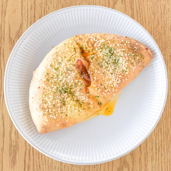 Golden-brown calzone with sesame seeds and herbs, oozing cheese and sauce, on a white plate.