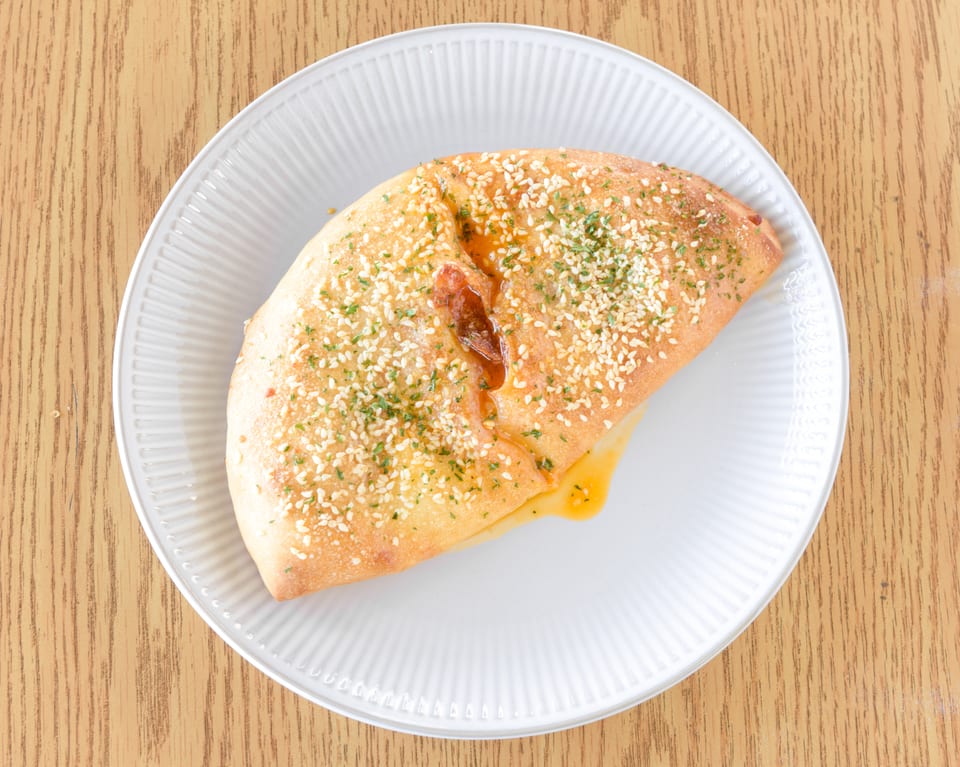 Golden-brown calzone with sesame seeds and herbs, oozing cheese and sauce, on a white plate.